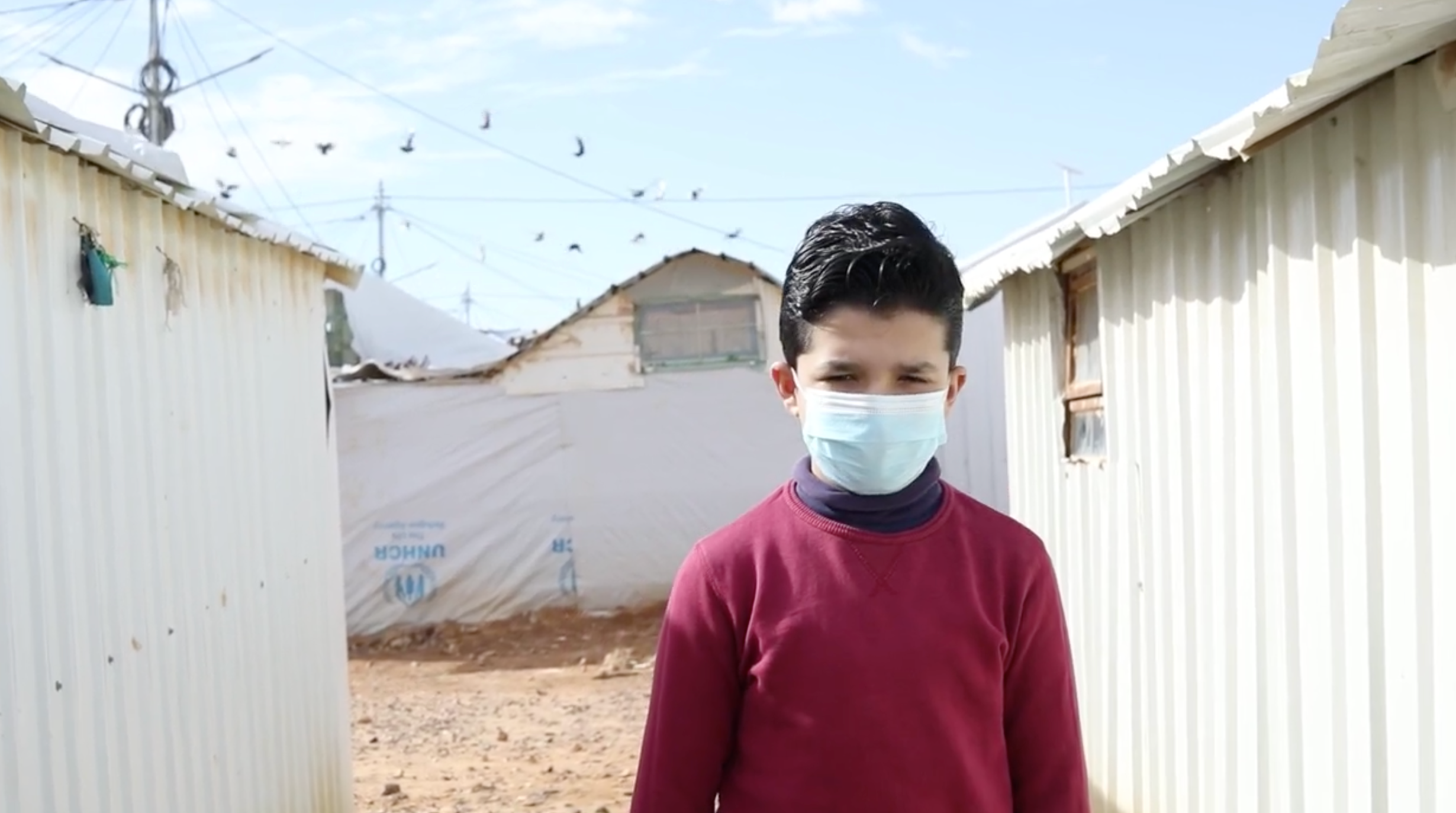 A boy wearing a surgical mask, surrounded by white makeshift houses in a refugee camp.