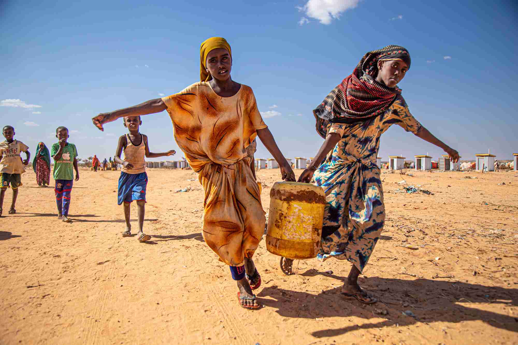 Two girls work together to carry a water canister over dry ground.