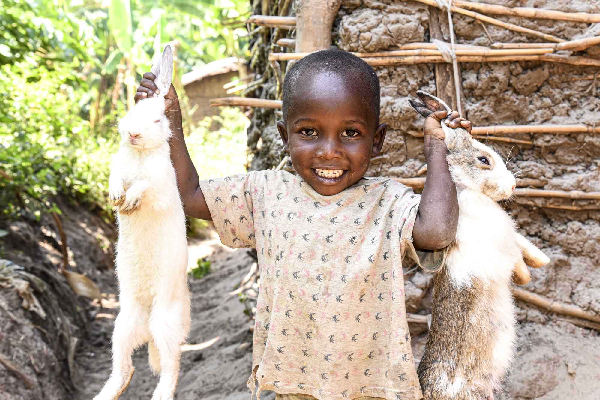 A young child stands outside smiling, while holding up two white rabbits by their ears.