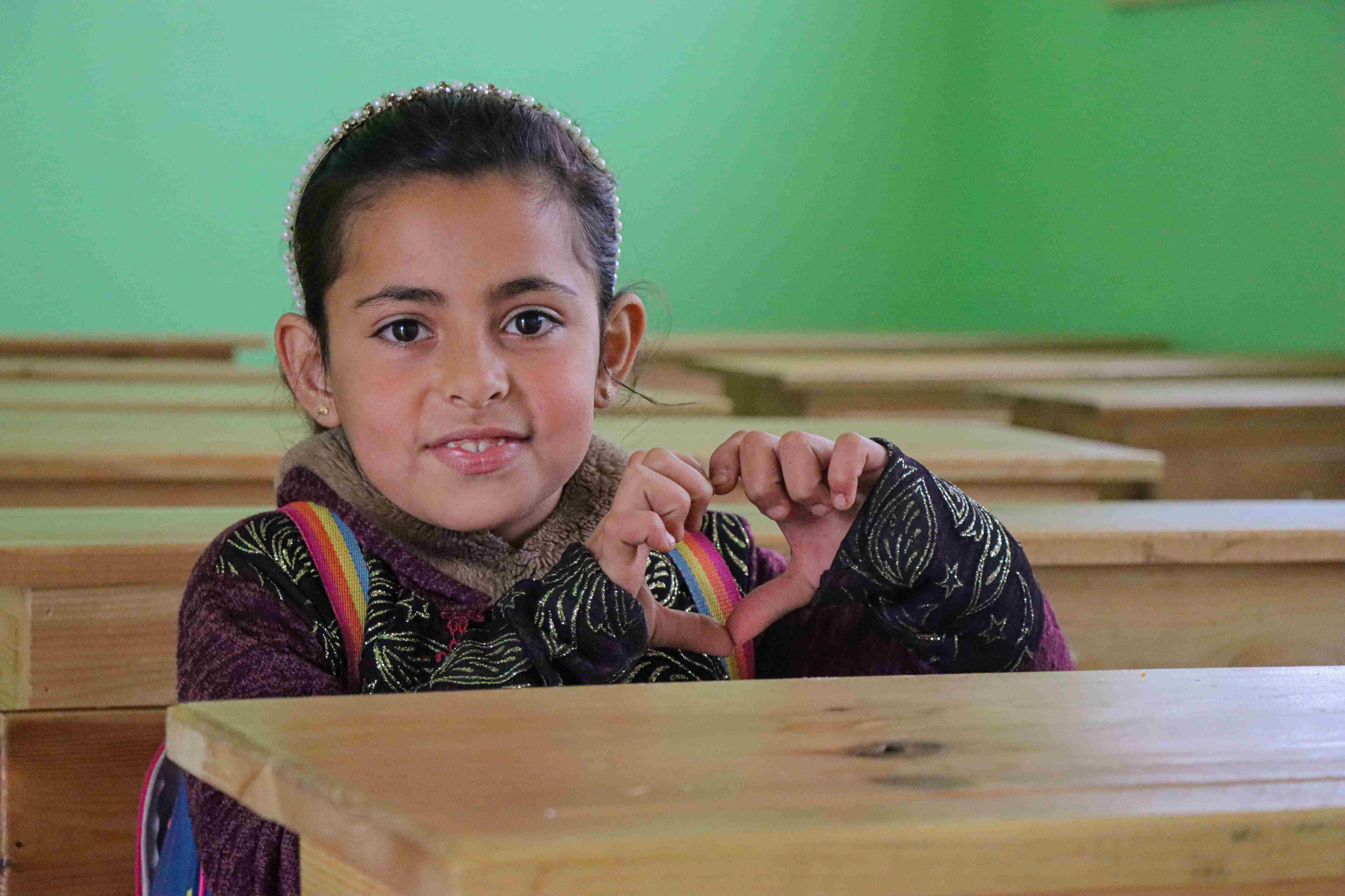 In Syria, a young girl smile and she sits at a school desk and makes a heart symbol with her hands.