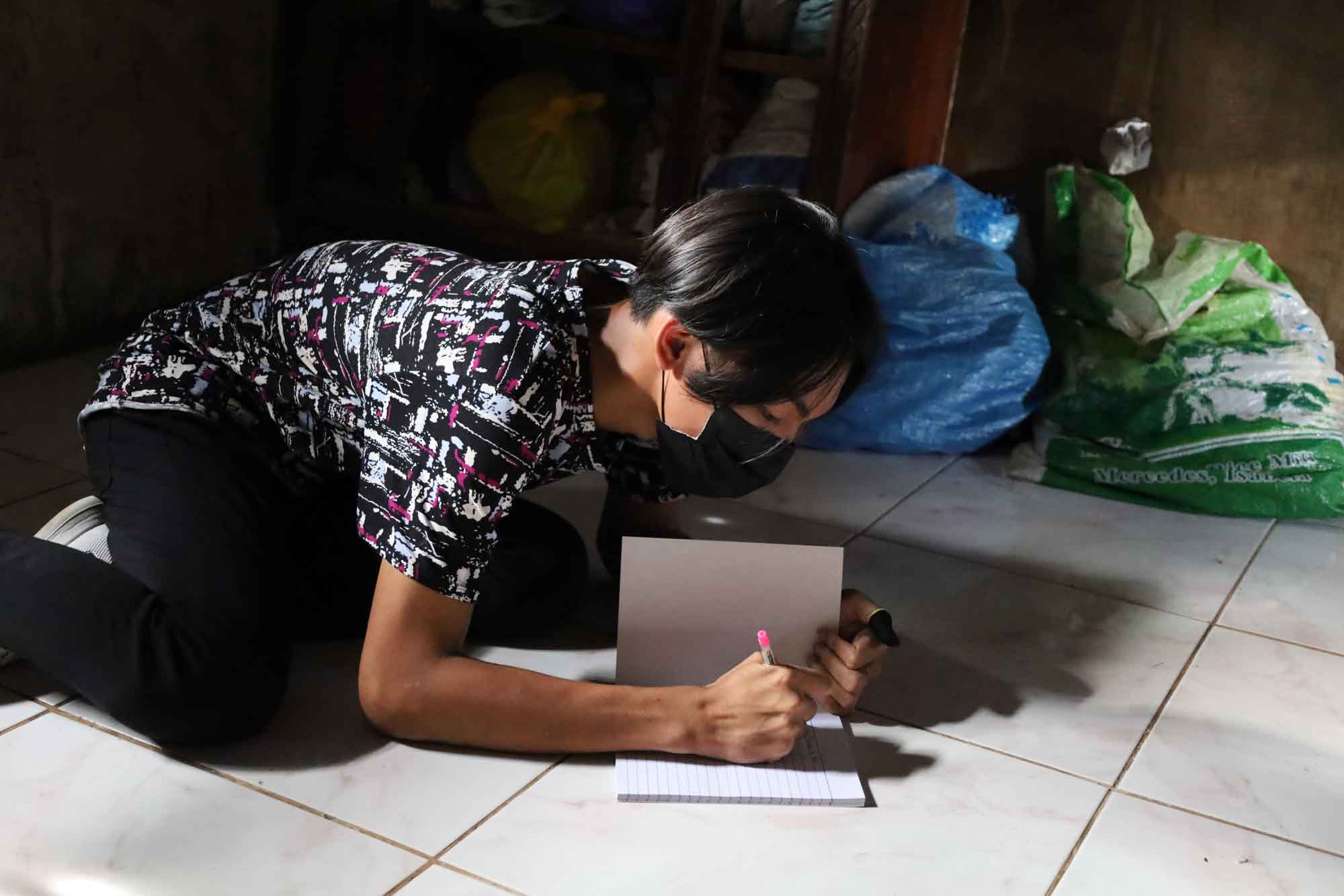 A person wearing a medical mask is seated on the ground, writing in a notebook.  