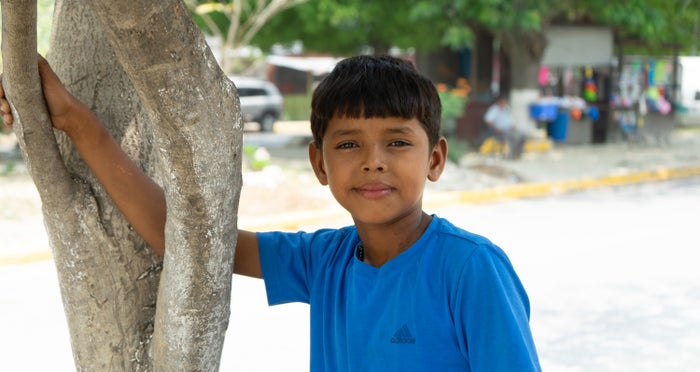 In Honduras a young boy leans against a tree with a closed-mouth smile. 