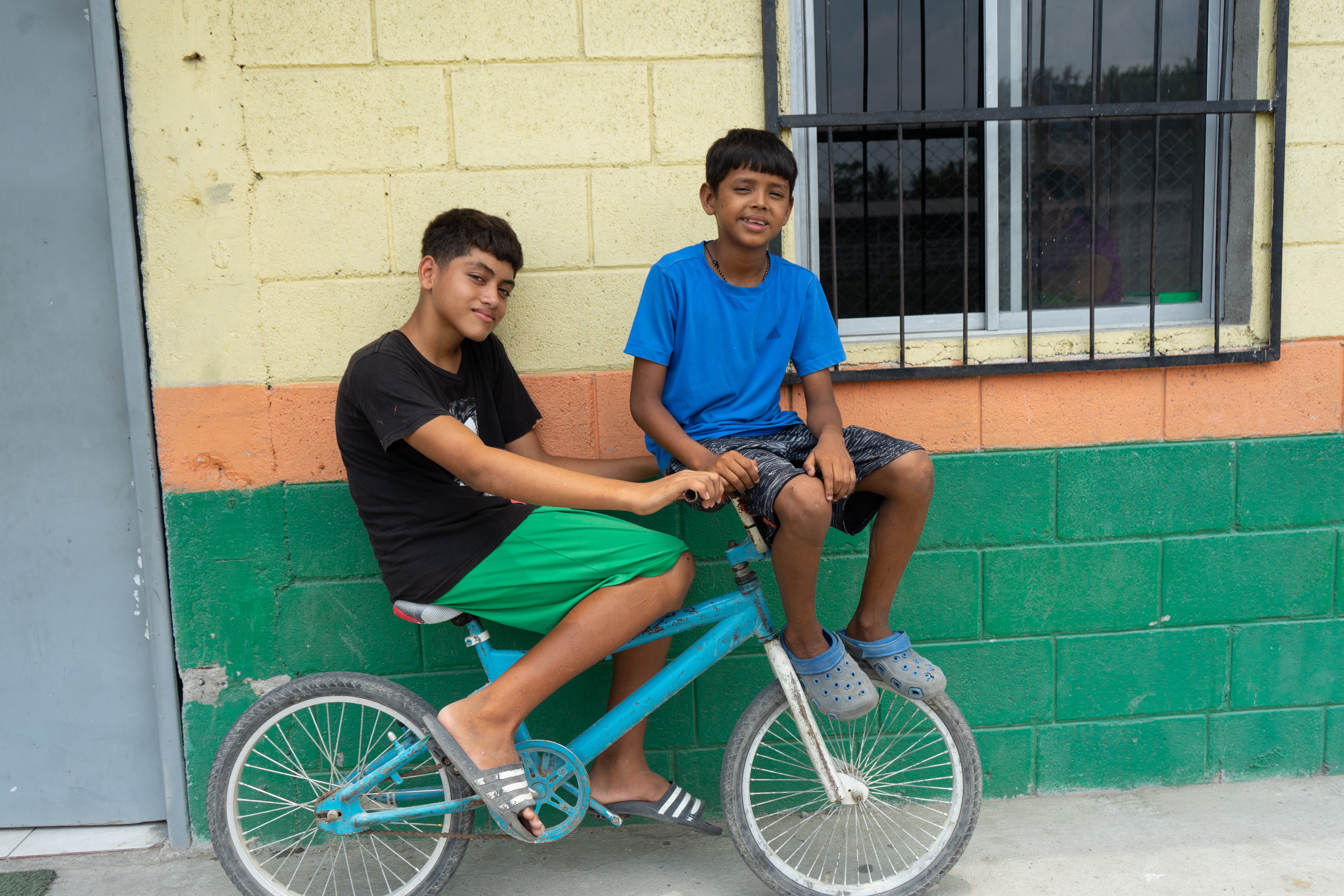 In Honduras, a young boy sits on his bike with his friend, Chulo sitting on the handle bars. A colorful wall in behind them. 
