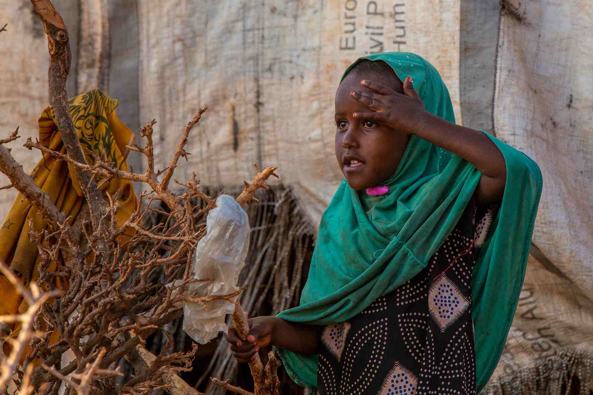 A young girl with a turquise head covering stands beside a makeshift shelter and shades her eyes from the sun.