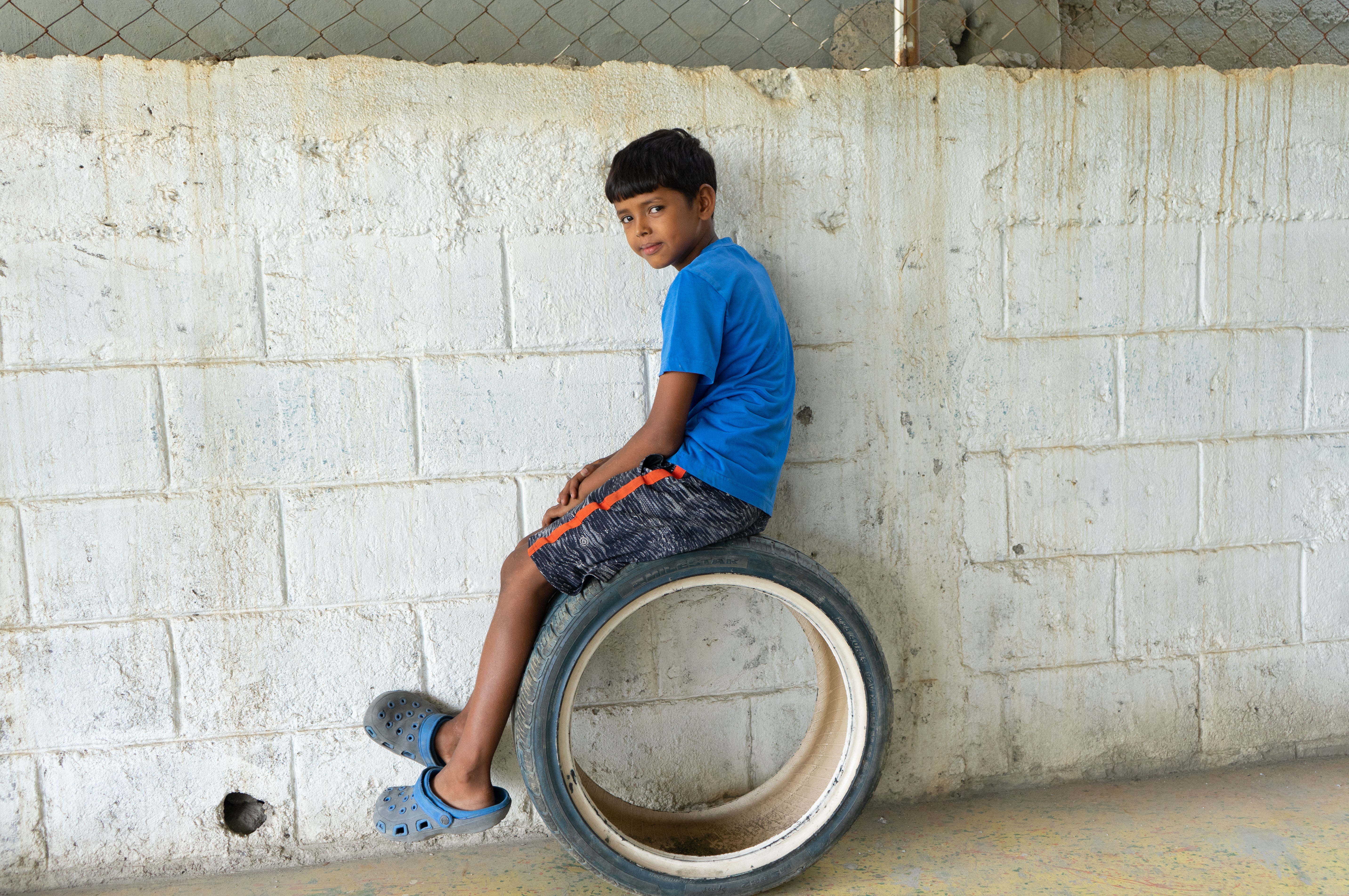 In Honduras a young boy sits on a tire against a white cement wall.  