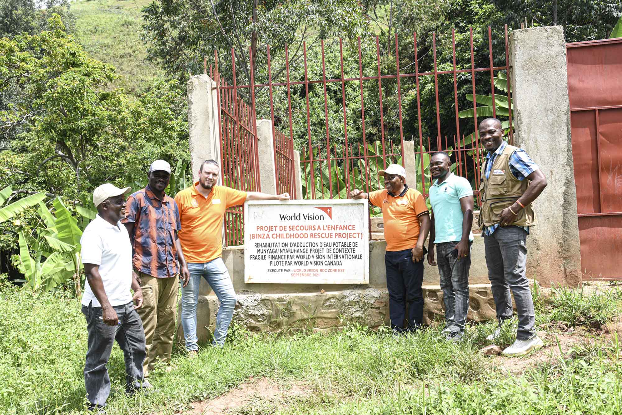 A group of men smile while standing together outdoors in front of a World Vision property sign.