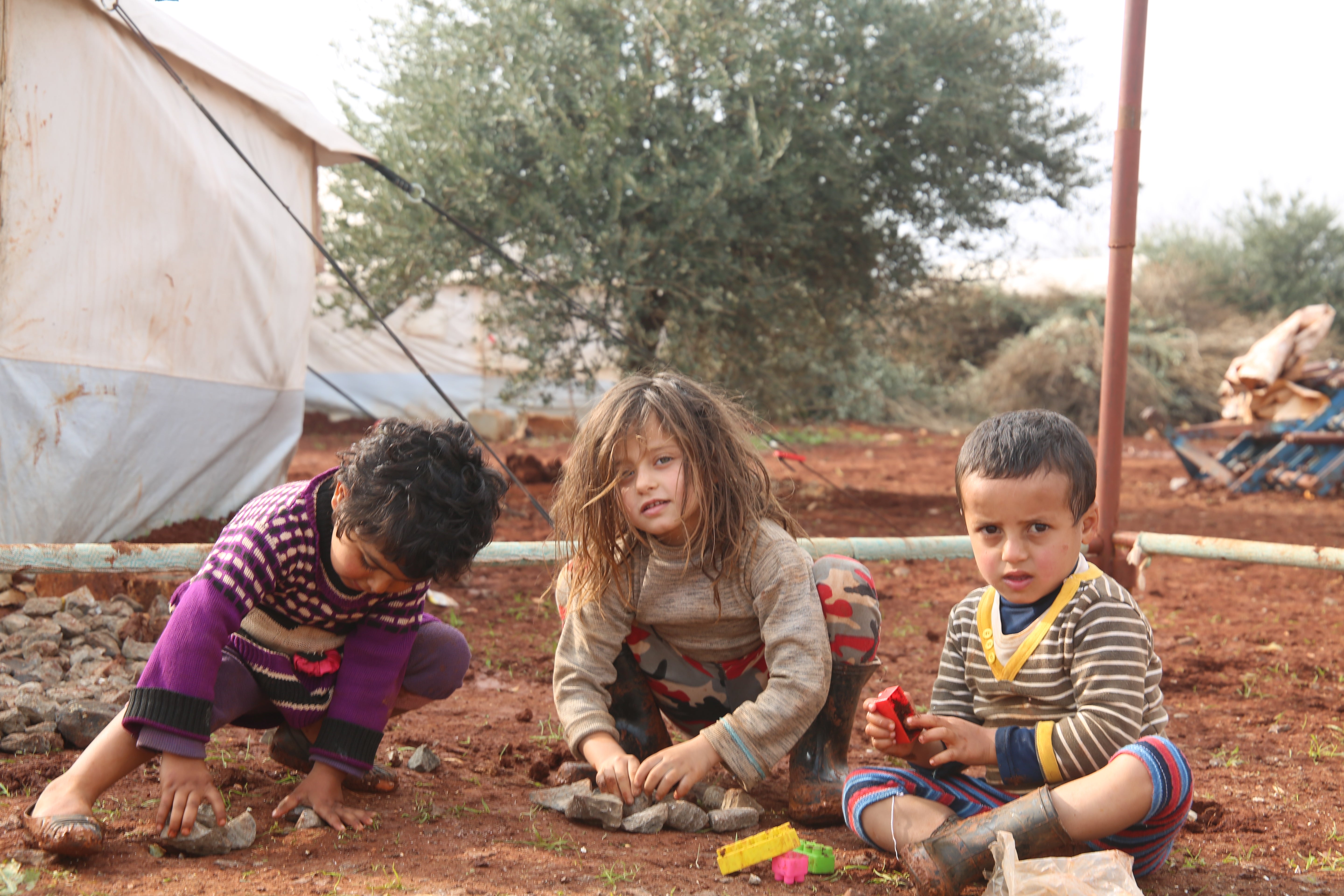 Three young children playing in the dirt outside their makeshift home.