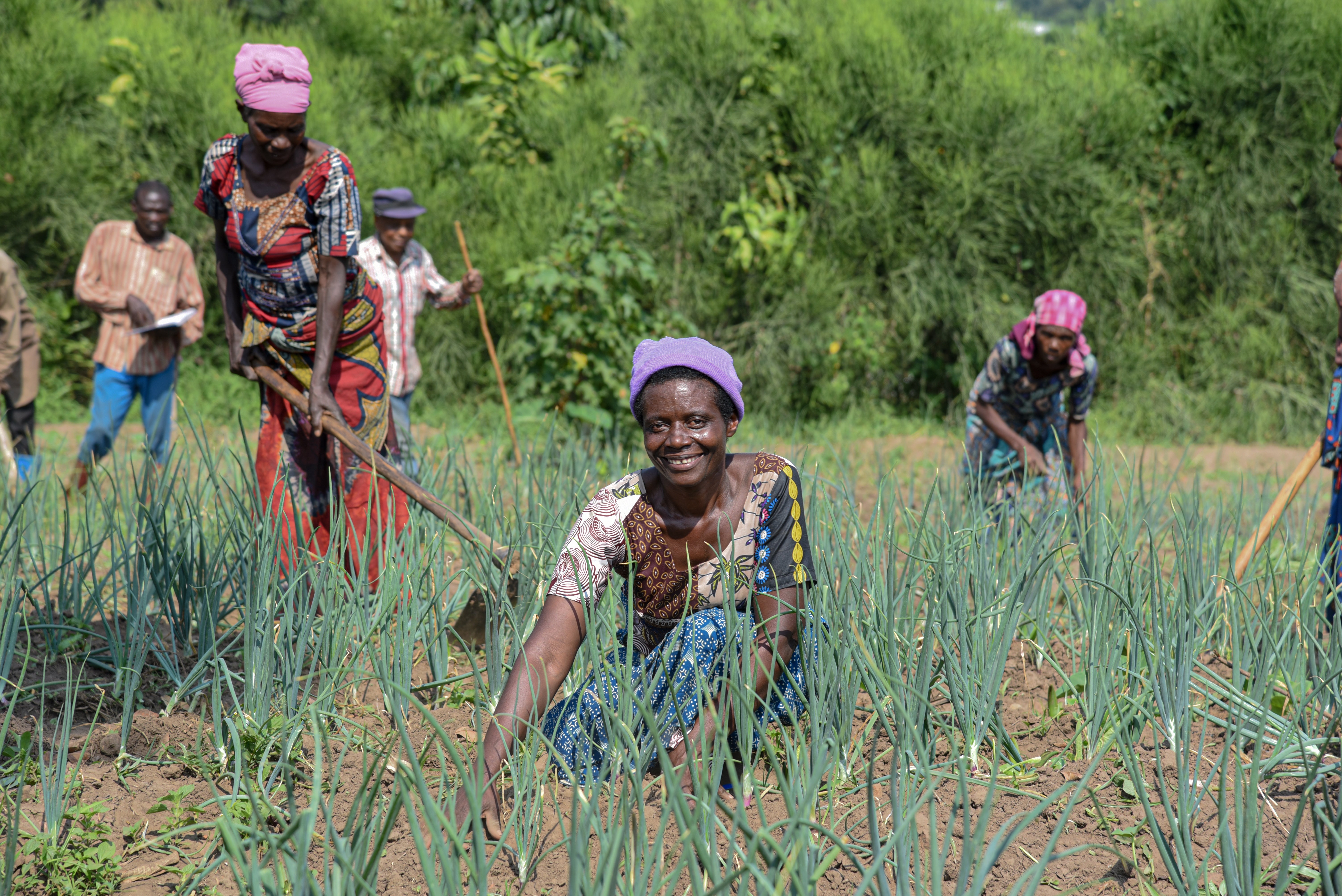 A women smiles as she kneels in her farm field. Behind her others work the fields.
