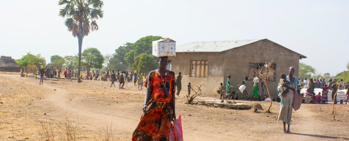 A woman standing outdoors with a white box on top of her head and a bag in her left hand.
