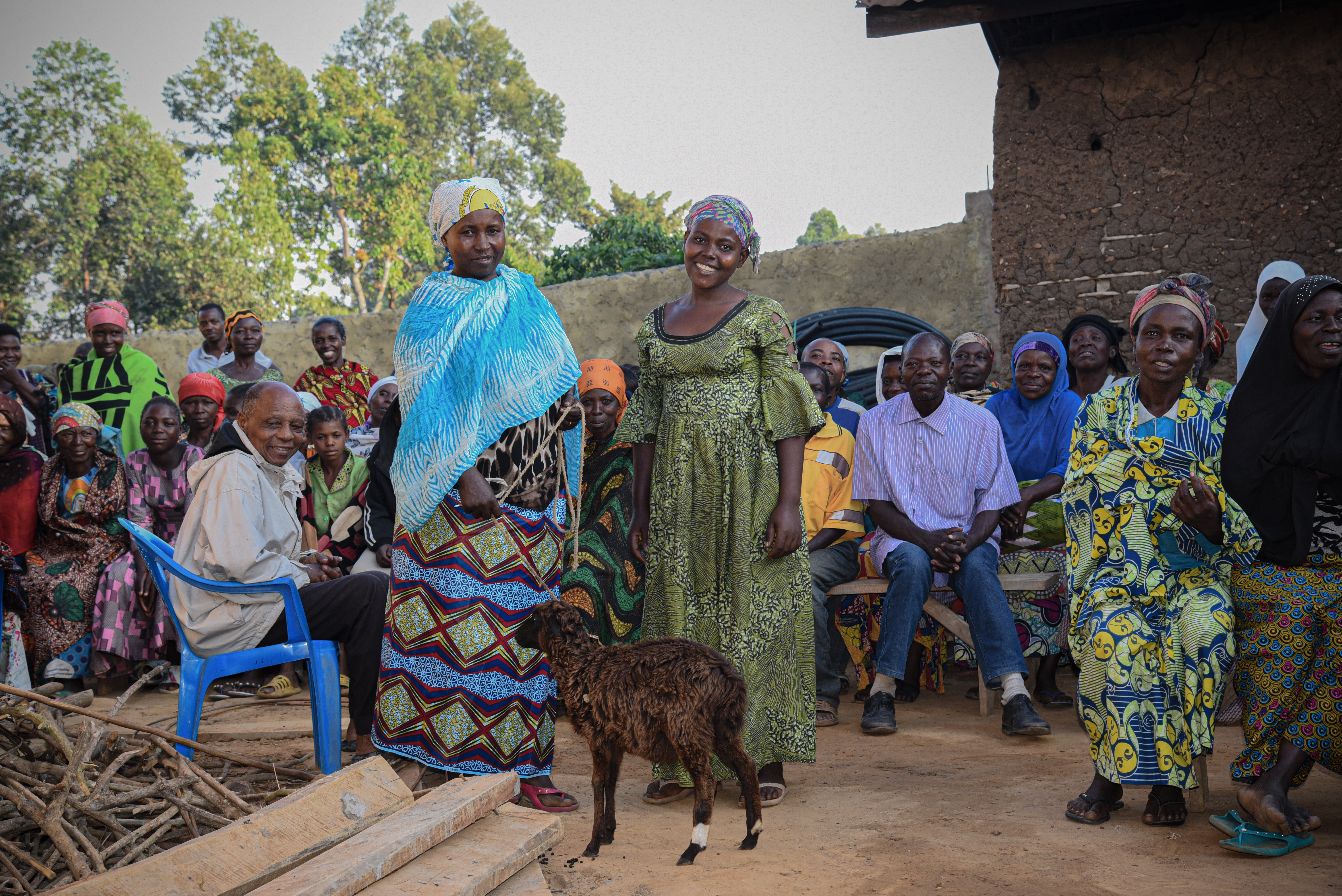 In the Democratic Republic of the Congo, two women smile and stand with a sheep between them. Other community members are around them.