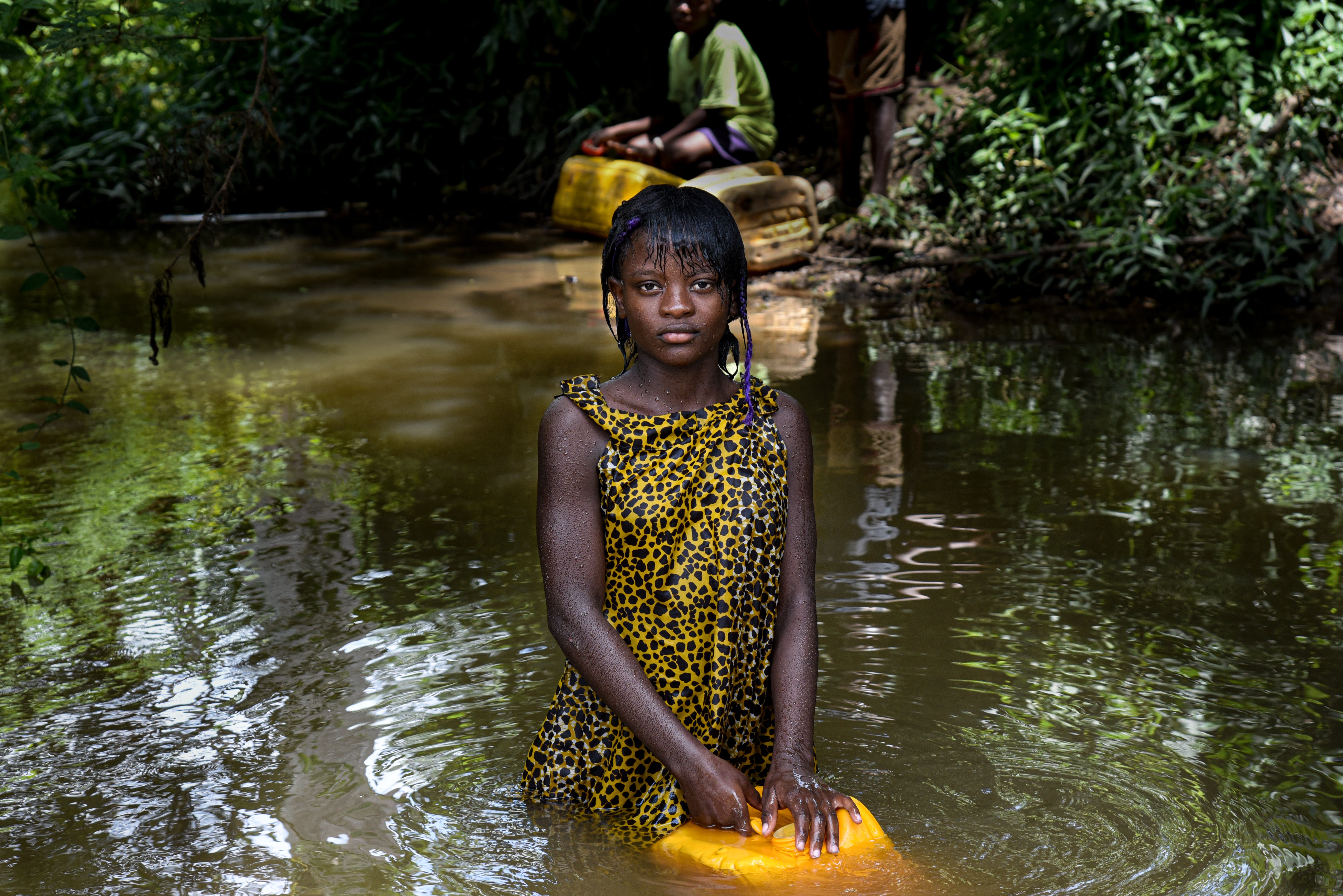 A young Congolese girl stands in a dirty river with a yellow dress on and looks at the camera. She holds a yellow water can and is filling it up.