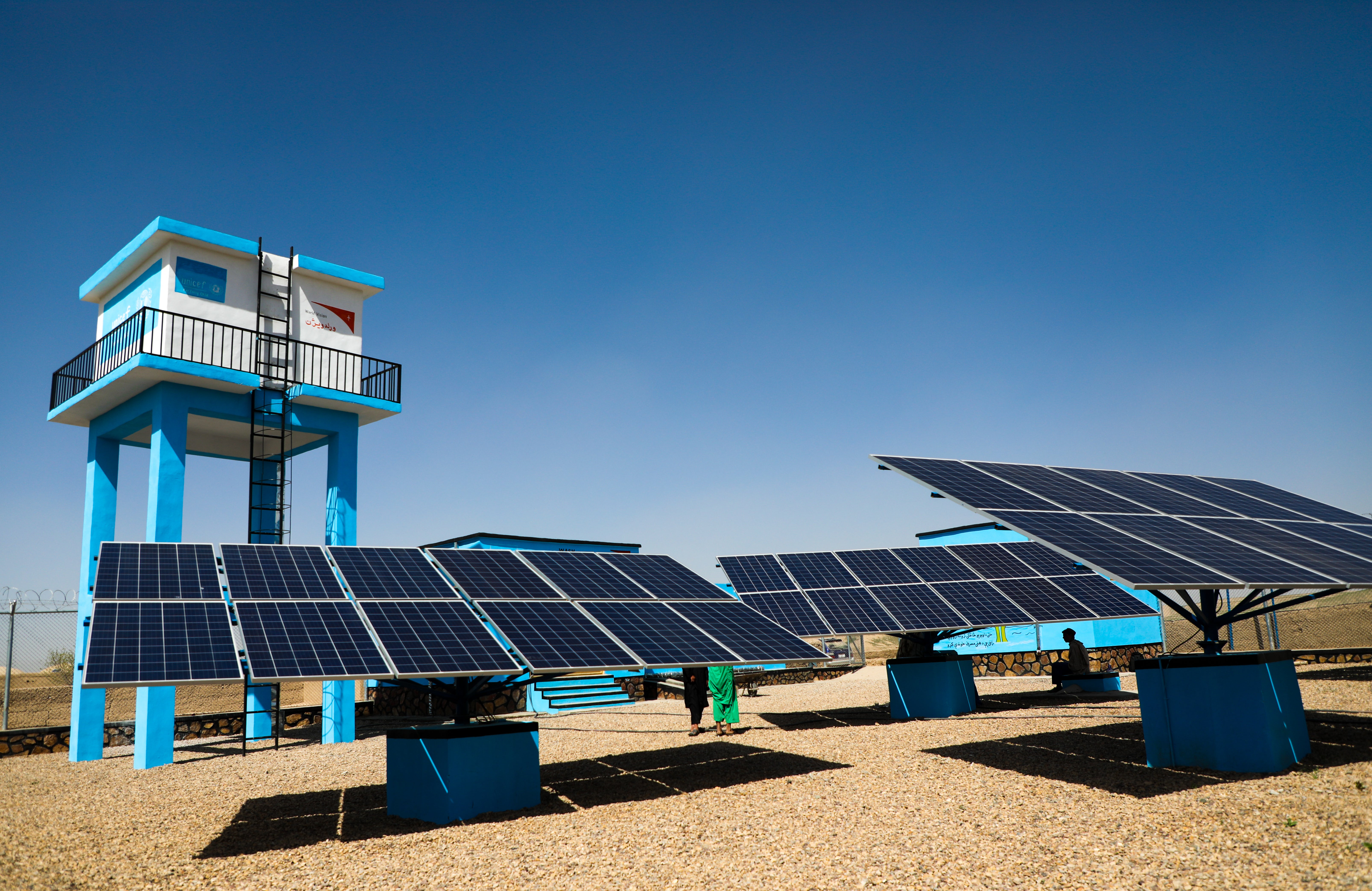 In Afghanistan large solar panels point towards the sky in a desert area. Behind the solar panels in a solar powered water station. 