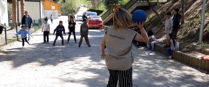A female World Vision staff playing dodgeball with a group of children on the street.