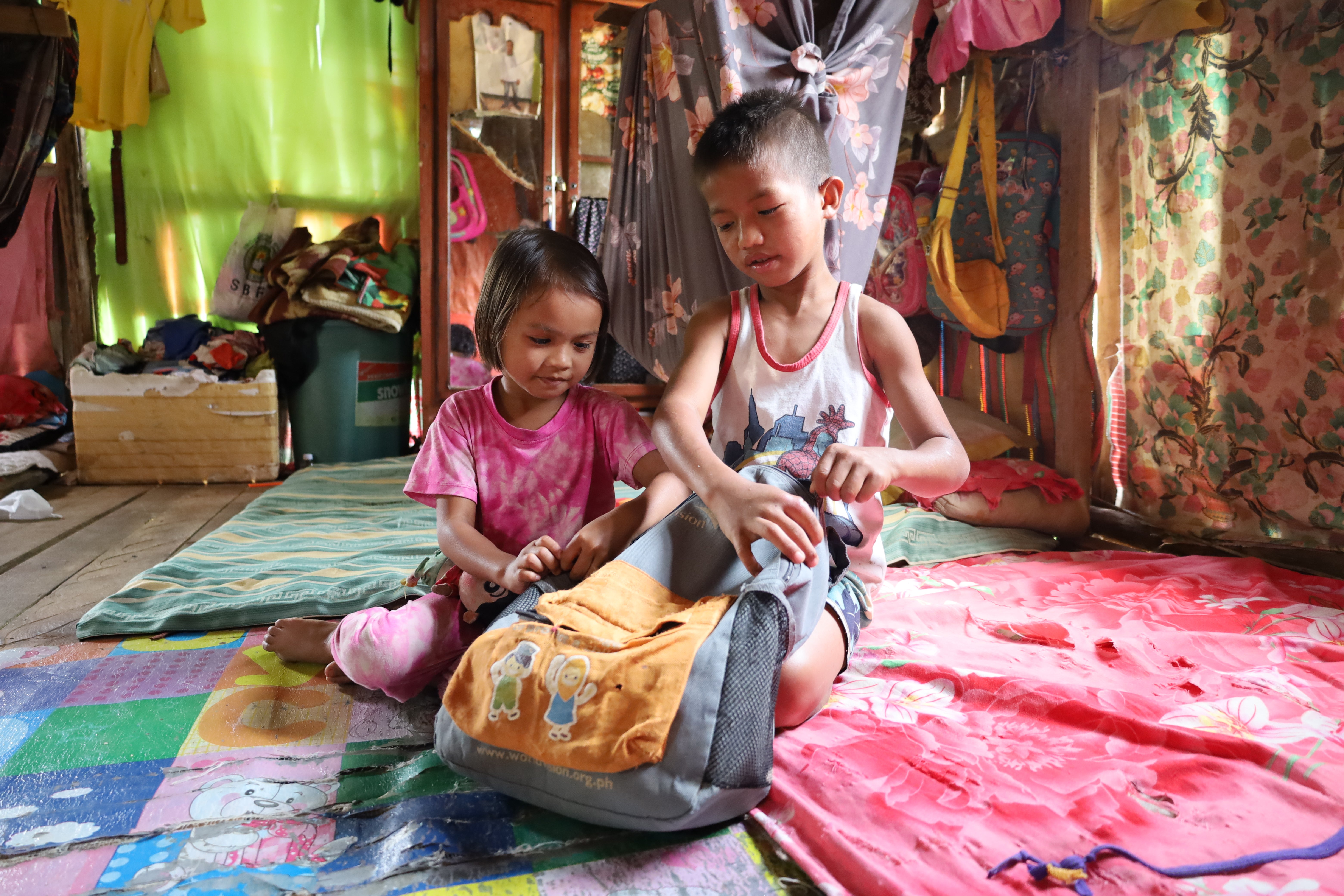 In the Philippines, a young boy sits with his younger sister and closes his backpack. 