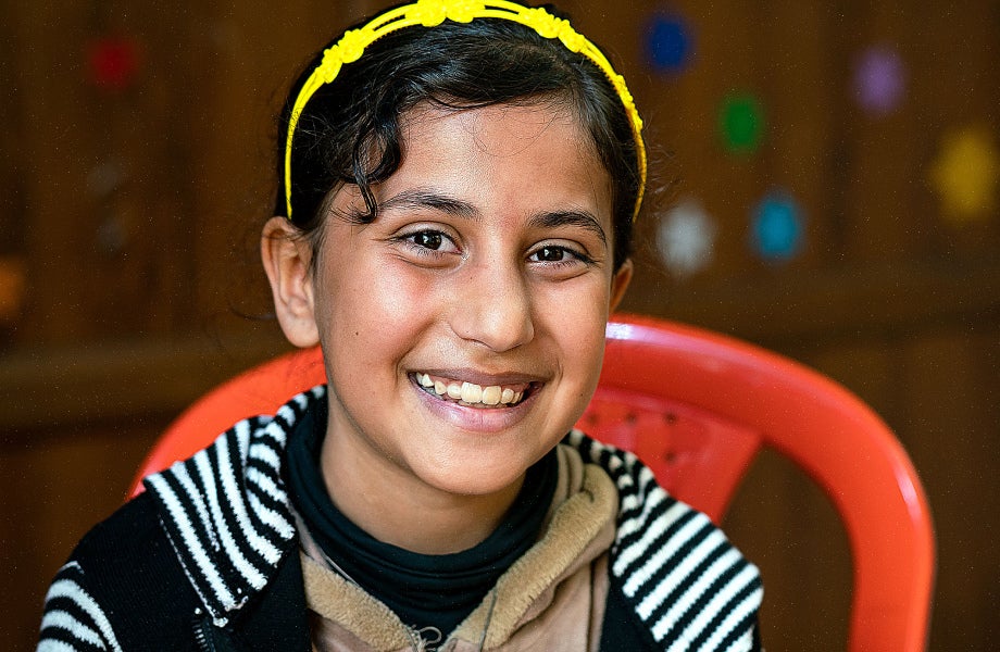 A girl wearing a bright yellow headband and sitting on a red monobloc chair, smiling happily.