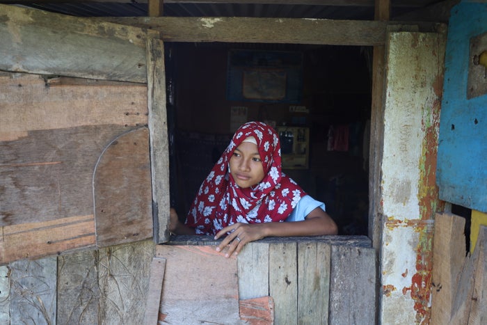 A young girl with a red floral head covering looks out the window of a wooden makeshift home. 