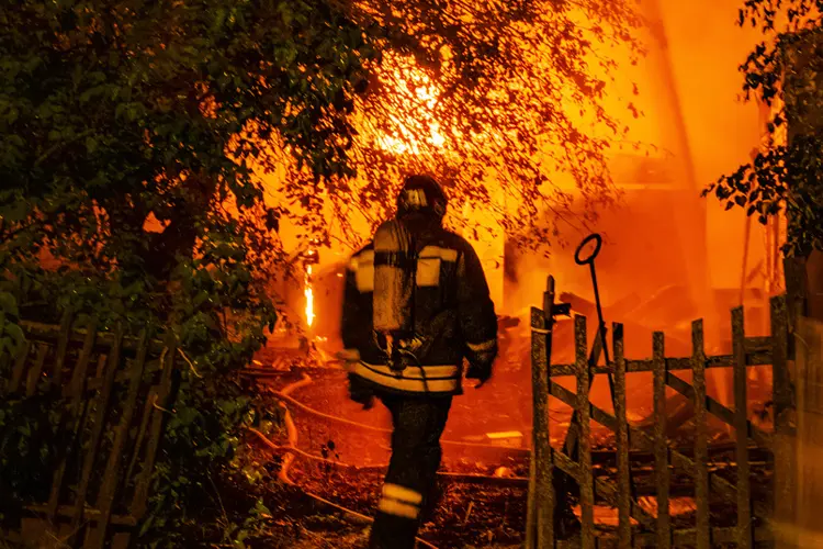 Firefighter silhouette against background of orange glow from fire in country house at night. If There's an Electric Fire in Your New House, Who Pays?