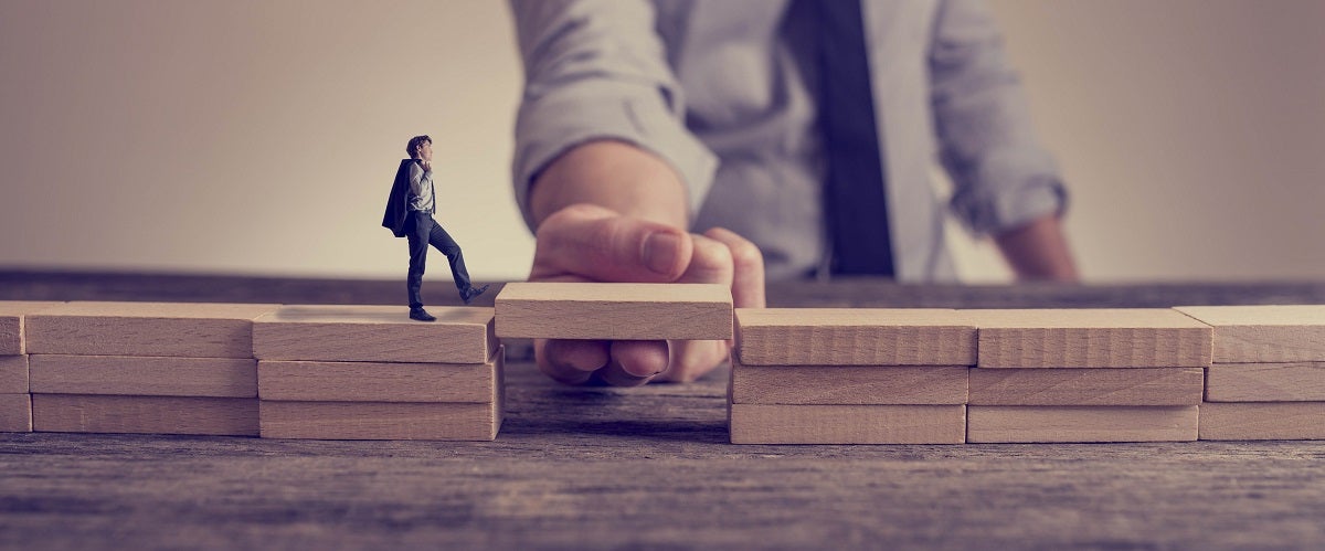 An image showing a man walking across a gap in building block using a block held by a large hand; a metaphor for the gaps in the use of GAAP for manufacturers.