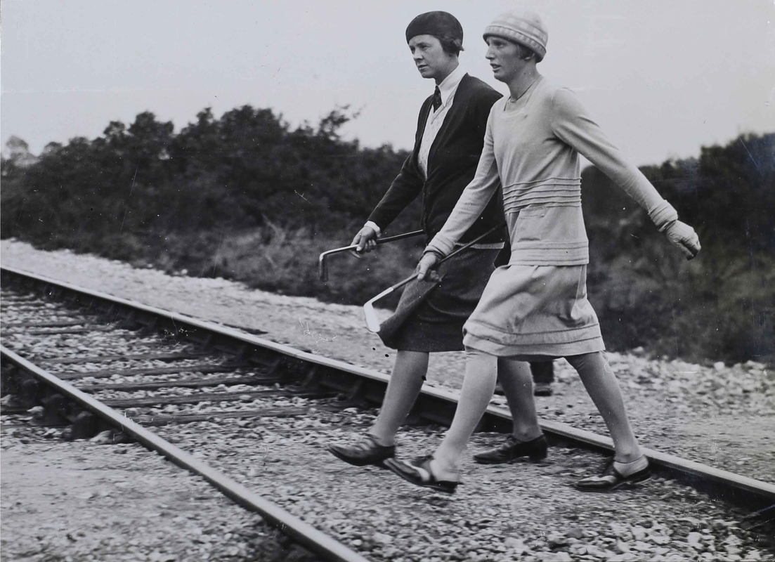 Two 1930s golfers, putters in hand, walking across a railway line.