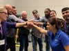 A group of training participants form a close-knit circle with their hands stacked in the center. They are mid-yell, faces animated with enthusiasm and unity, embodying teamwork and camaraderie.