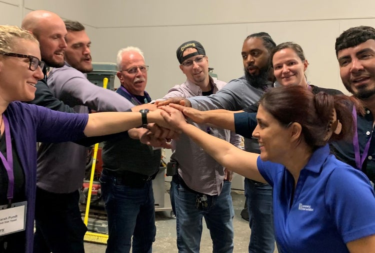 A group of training participants form a close-knit circle with their hands stacked in the center. They are mid-yell, faces animated with enthusiasm and unity, embodying teamwork and camaraderie.