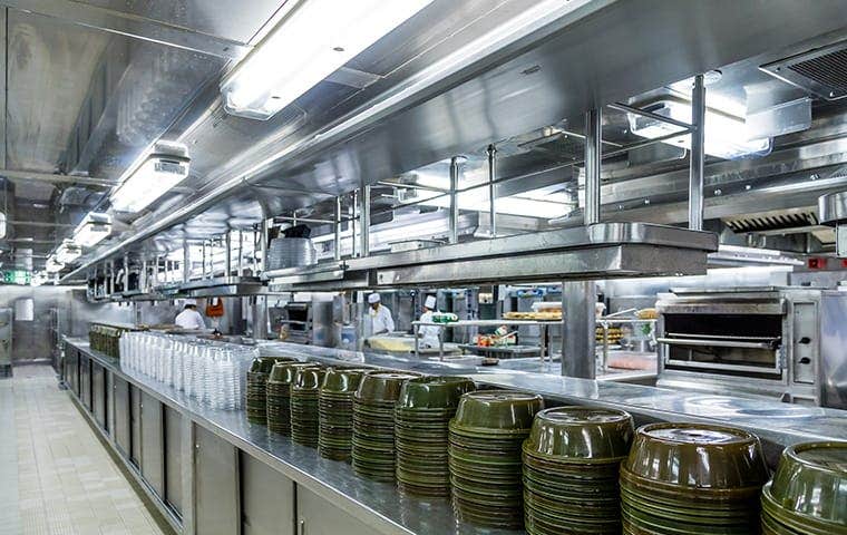 Inside of an industrial commercial kitchen. Full of stainless steel appliances at a larger scale, in an organized fashion, are green plates and to-go packages going down the prepping edge on the countertop.