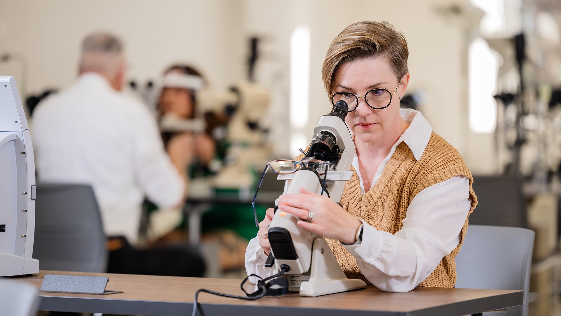 A female eyeglasses student measures lens power for a pair of eyeglasses using a manual lensometer. The course instructor in the background examines another female student's retina with a slit lamp refractor.