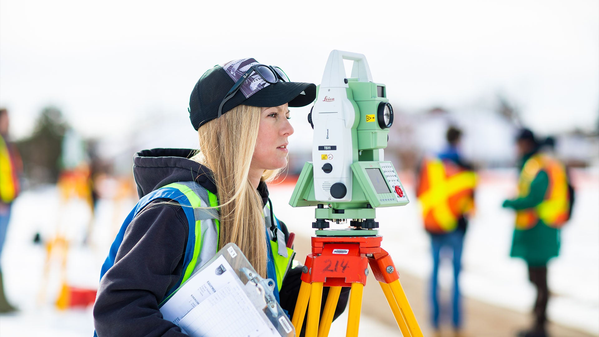 A female surveyor wearing a black cap and reflective vest operates a Leica TS11 total station on a tripod in an outdoor setting, holding a clipboard.