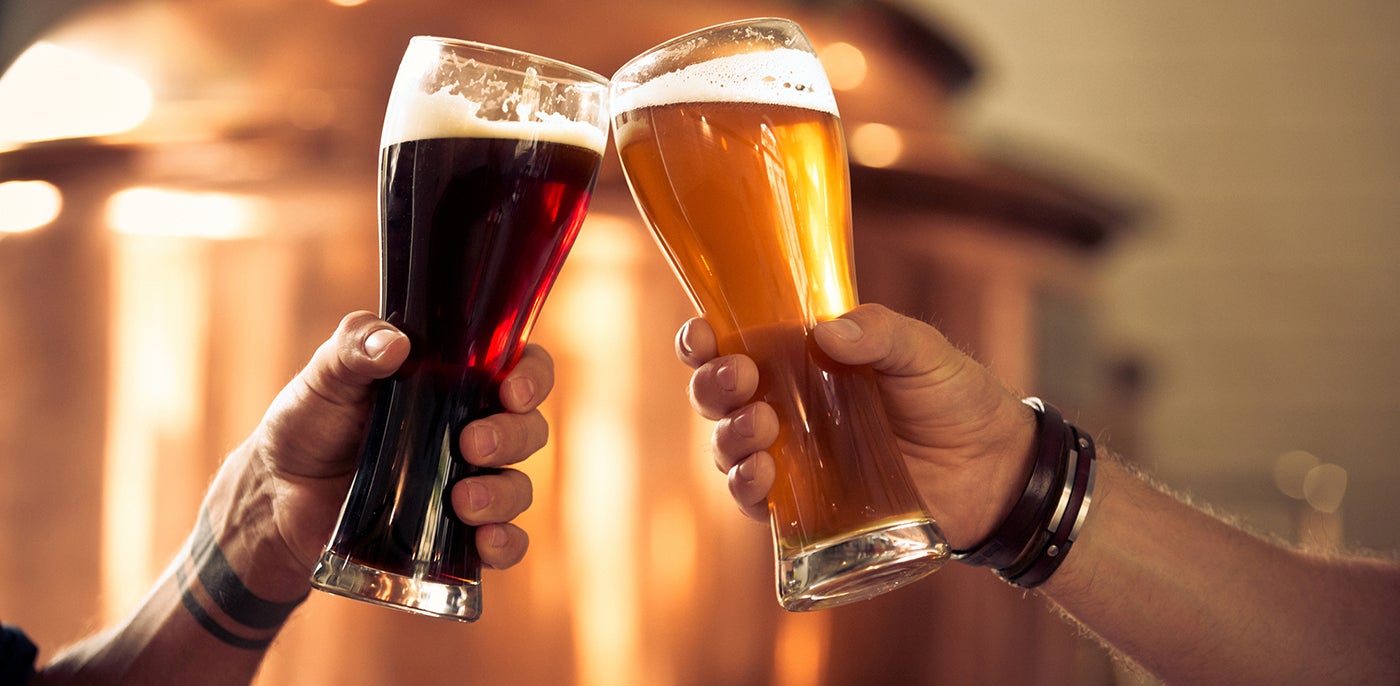 Two hands clink tall glasses of dark and amber beer, with a softly blurred brewery or pub setting in the background.