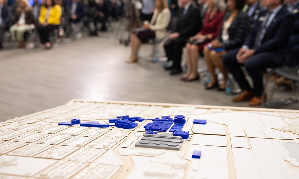 Scale model of a site layout with blue building blocks in the foreground and an audience seated and blurred in the background.