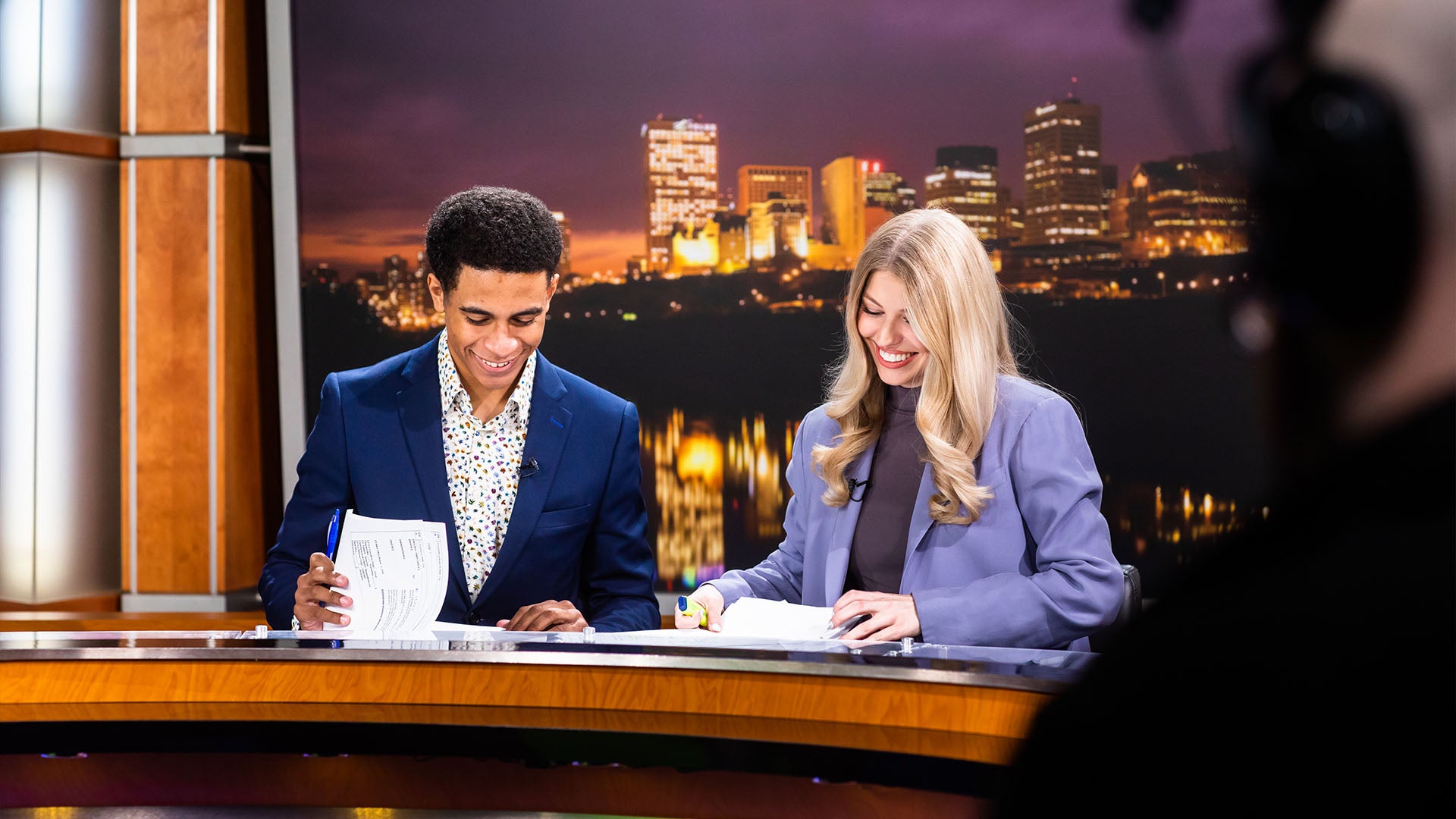 Two formally dressed students sit at a desk in the NAIT News Watch broadcasting studio.