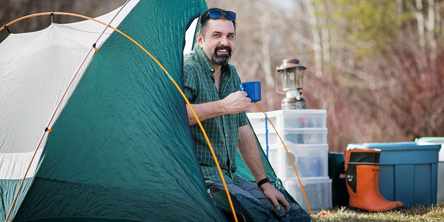 Person seated at the entrance of a camping tent holding a mug, surrounded by camping gear, storage bins, and a lantern in an outdoor campsite.