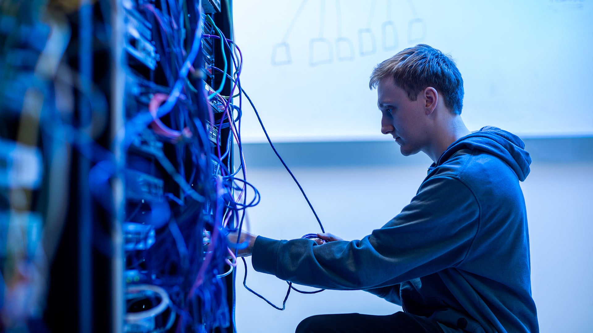 Man in a server room organizing and connecting cables, working on network switches, surrounded by colorful wires.