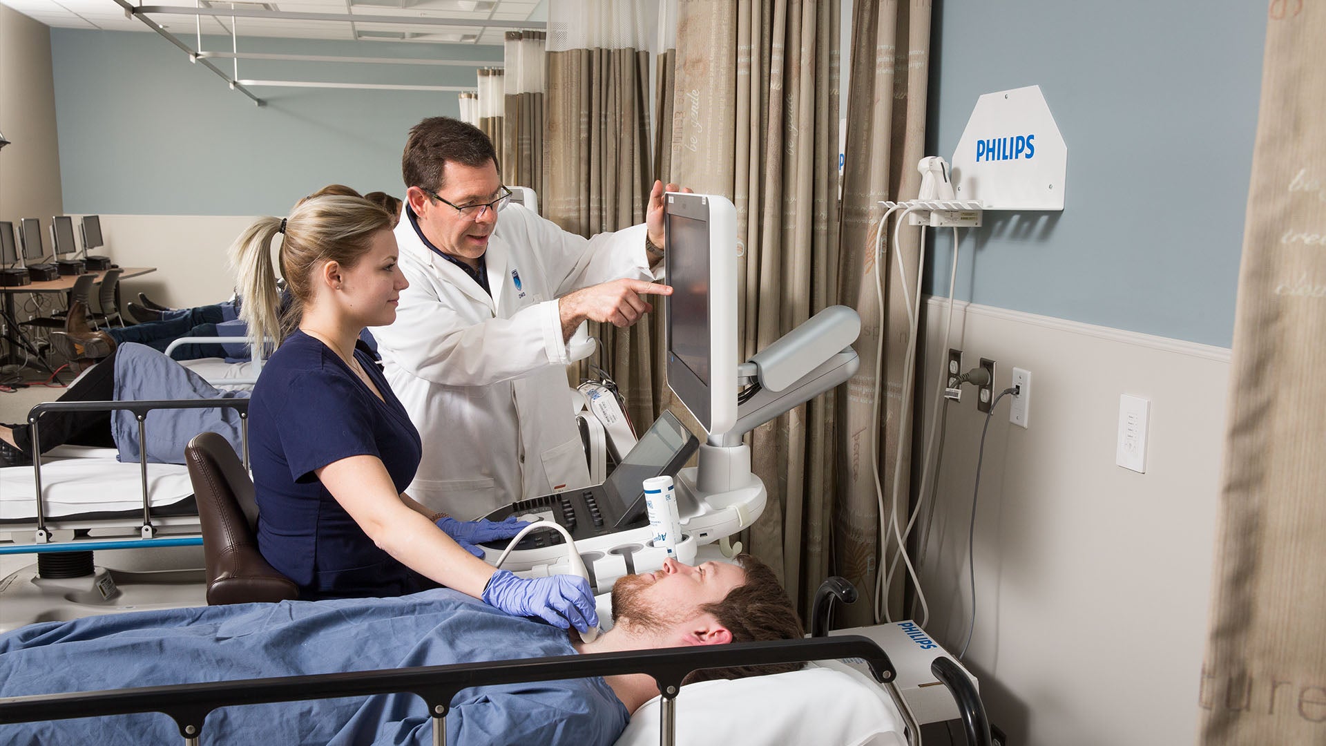 A sonographer looks at a monitor while scanning a patient's throat with an ultrasound probe. An instructor points at the screen.