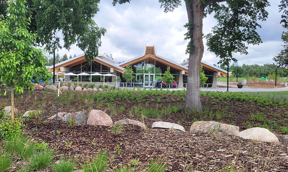 Modern park pavilion with a peaked wood-and-glass roof, viewed across landscaped gardens with trees, rocks, and mulched planting beds under a cloudy sky.