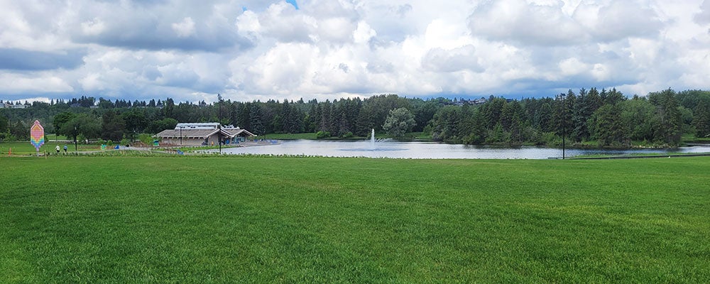 Wide view of a park lawn overlooking a lake with a fountain, surrounded by trees and a pavilion under a cloudy sky.