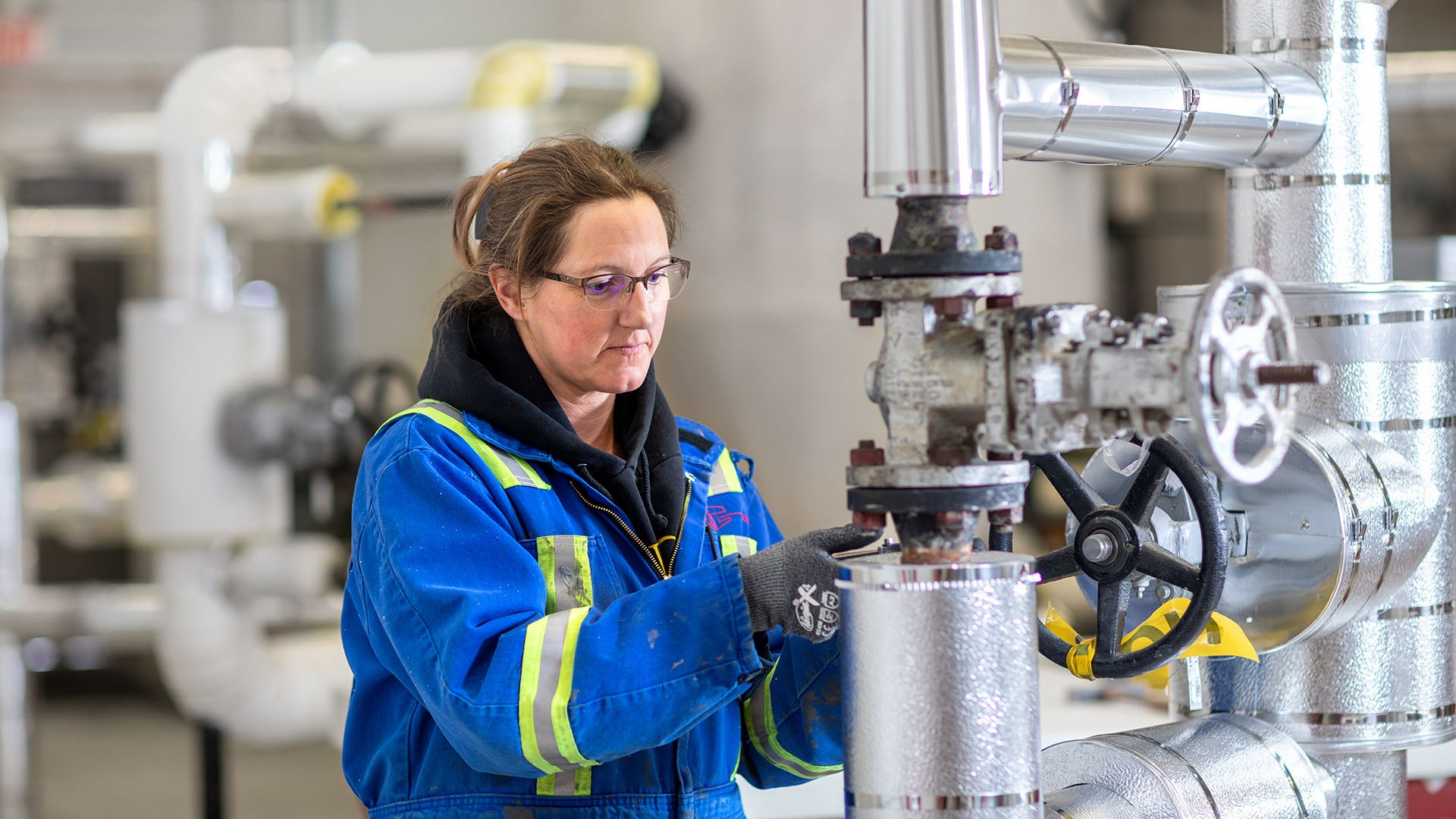 A female insulator in coveralls stands beside piping, touching a section covered with embossed metal jacketing over insulation, cladding she has installed or is inspecting. A female insulator in coveralls stands beside piping, touching a section covered with embossed metal jacketing over insulation, cladding she has installed or is inspecting.