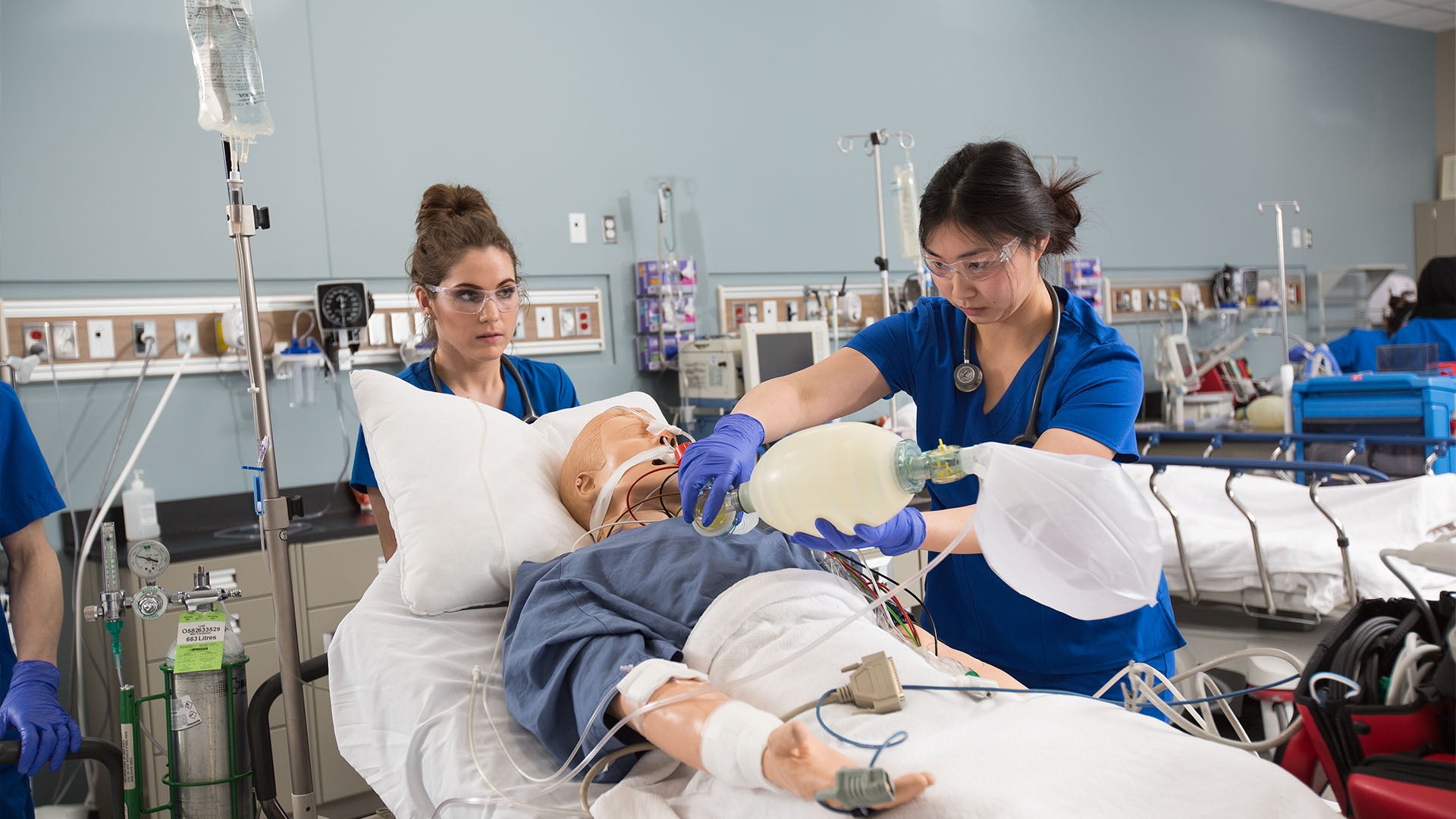 A respiratory therapist is wearing protective glasses and holding a breathing tube while looking at the patient. Another therapist is observing from the back.