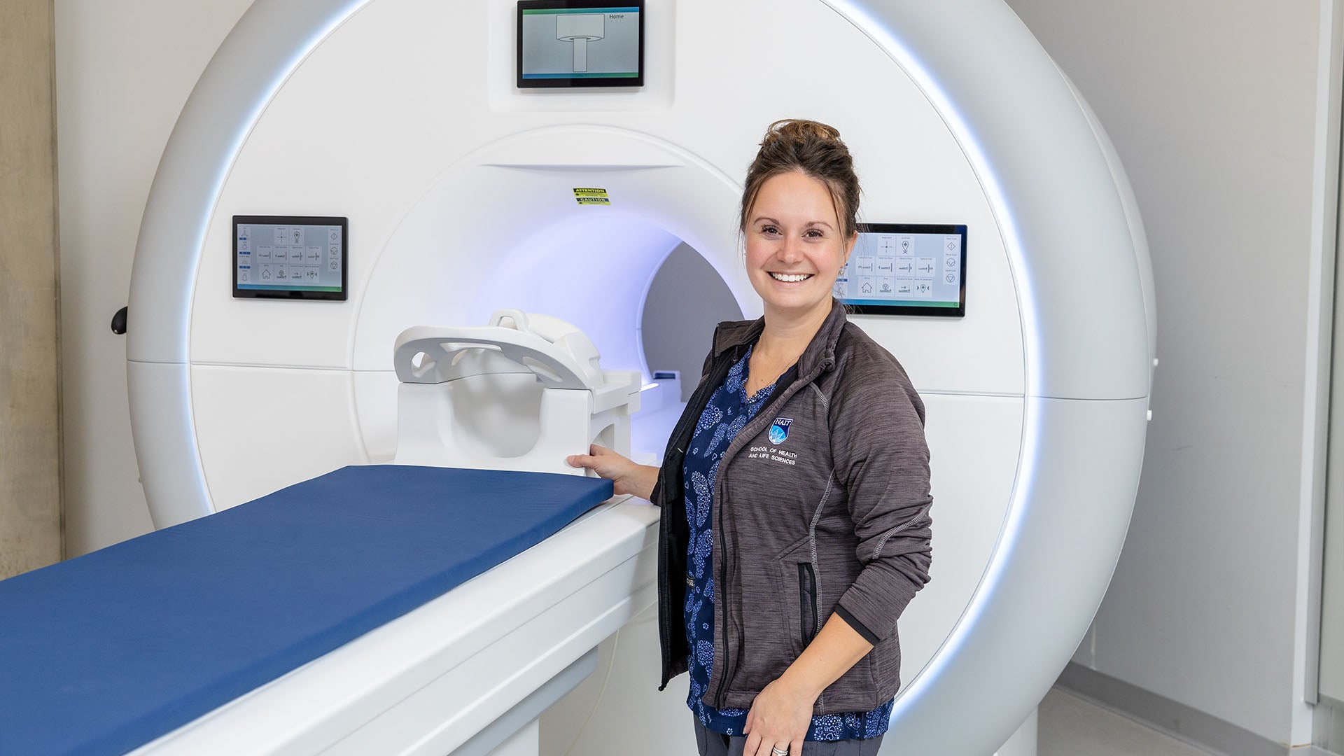A woman wearing a jacket with NAIT branding smiles while standing next to an MRI machine.