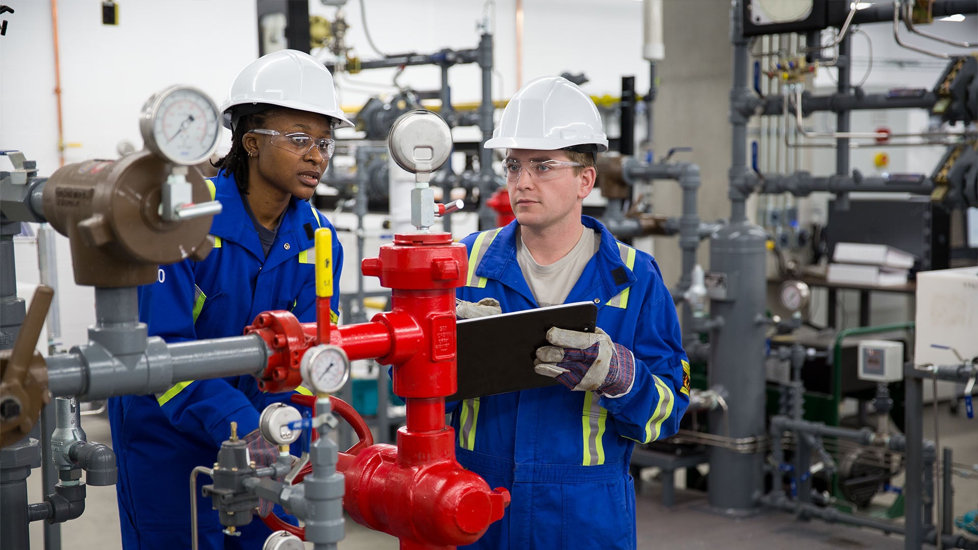 Two students in blue coveralls, hard hats, and safety glasses work together in an industrial lab. One holds a clipboard while the other operates complex piping and machinery.