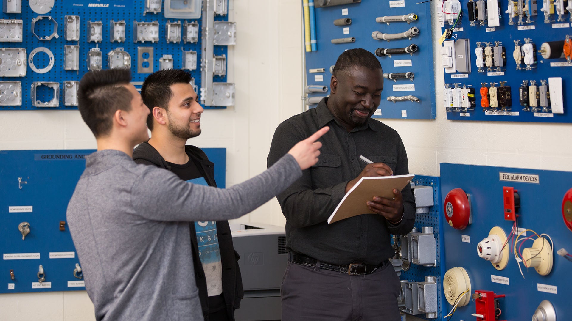 Two electrical engineering students talking to their instructor while he writes on a notepad.