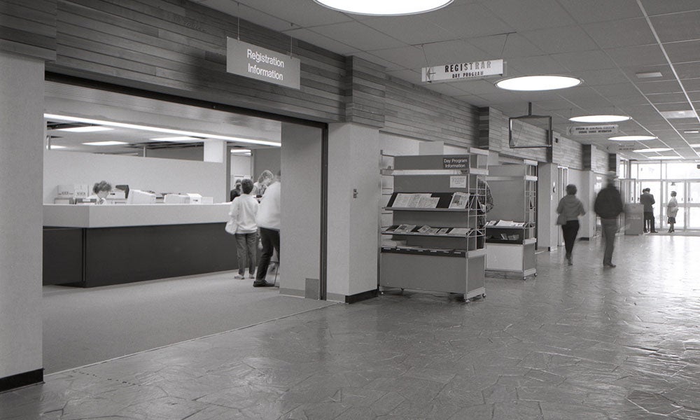 NAIT service and information counter area with brochure displays, overhead directional signage, and people walking through a wide interior corridor.