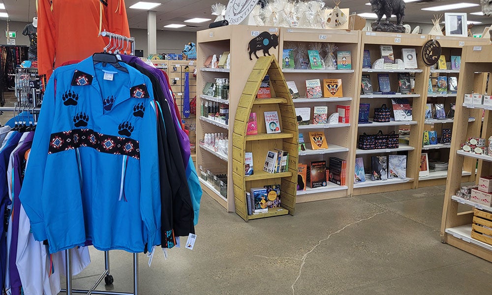 Bright blue Indigenous-style pullover with floral and paw-print designs displayed in a gift shop aisle lined with books and local merchandise.