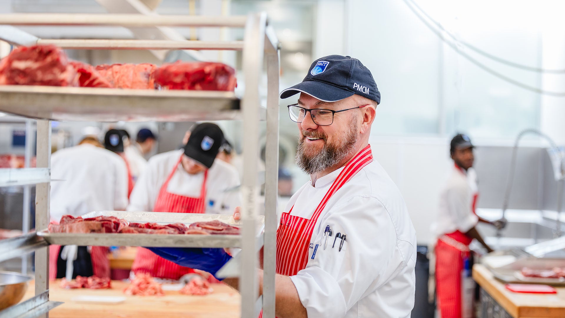 A smiling student wearing glasses, a baseball cap, and a striped apron carefully places a tray of sliced meat onto a storage rack.
