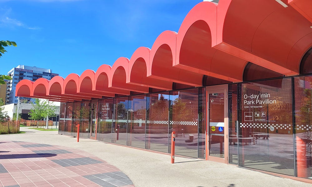 Modern glass-walled park pavilion with a distinctive red scalloped roof canopy, situated in an urban plaza surrounded by trees and downtown buildings under a clear blue sky.