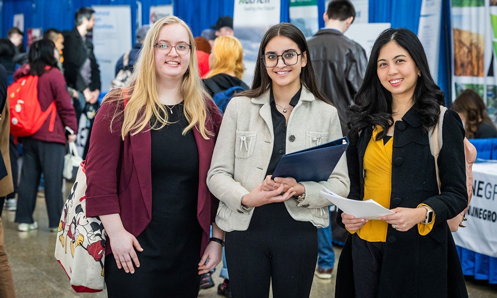 Three students standing together at a campus event, holding documents and a folder, with information booths and attendees in the background.