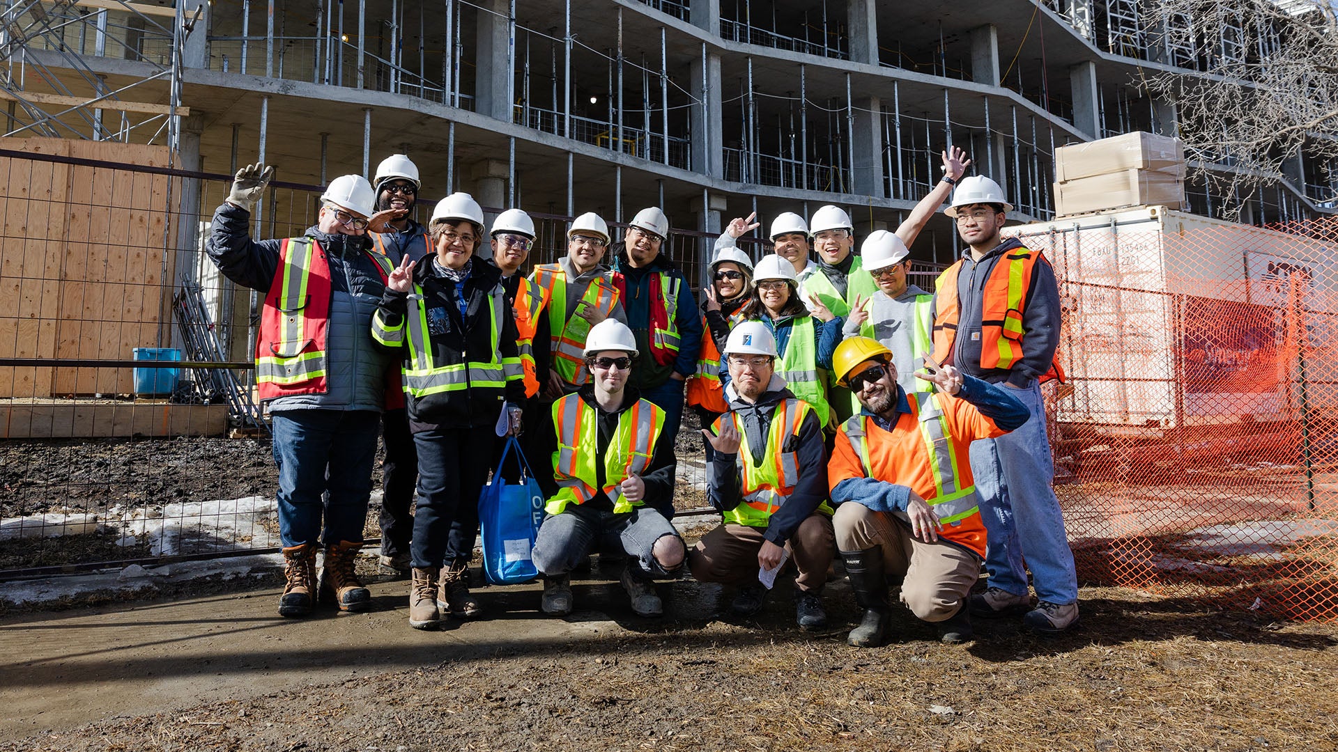Group of people wearing safety vests, hard hats, smiling and posing in front of a building under construction with scaffolding and fencing around the site.
