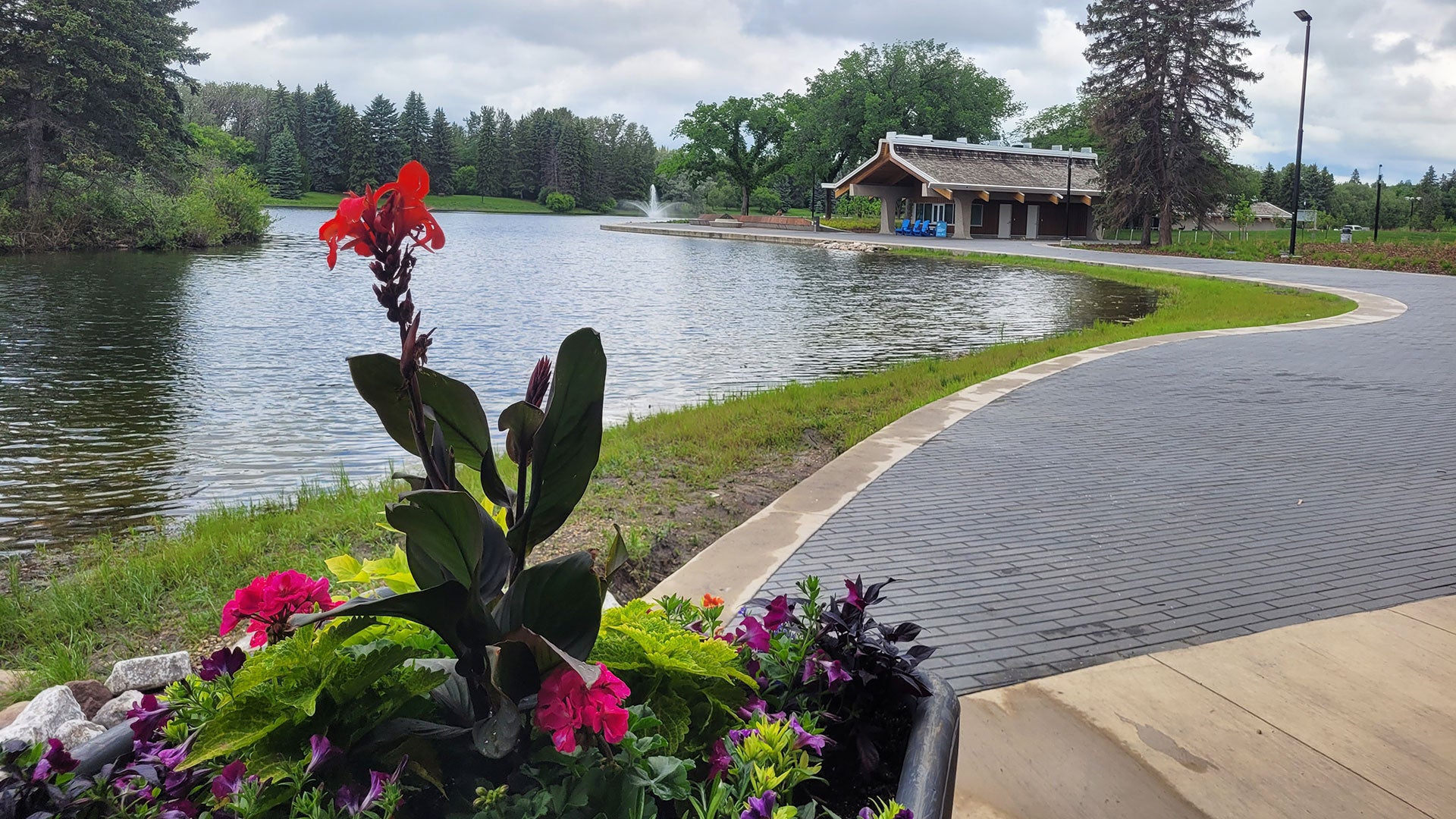 Colorful flowers in a planter beside a lakeside walkway, with a fountain, trees, and a pavilion under a cloudy sky. Colorful flowers in a planter beside a lakeside walkway, with a fountain, trees, and a pavilion under a cloudy sky.