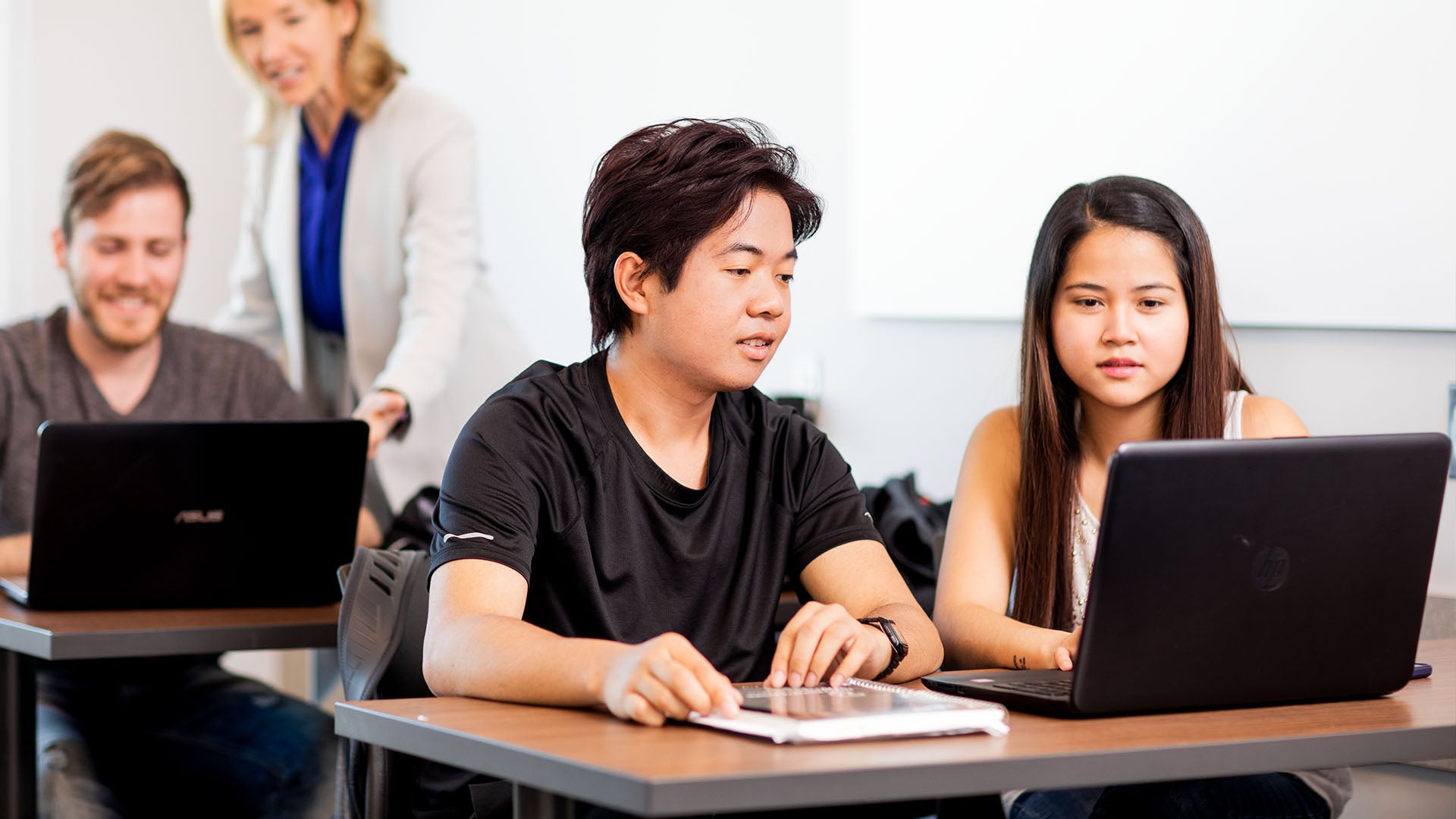 Two students looking at a laptop together, while an instructor helps another student seated behind them.
