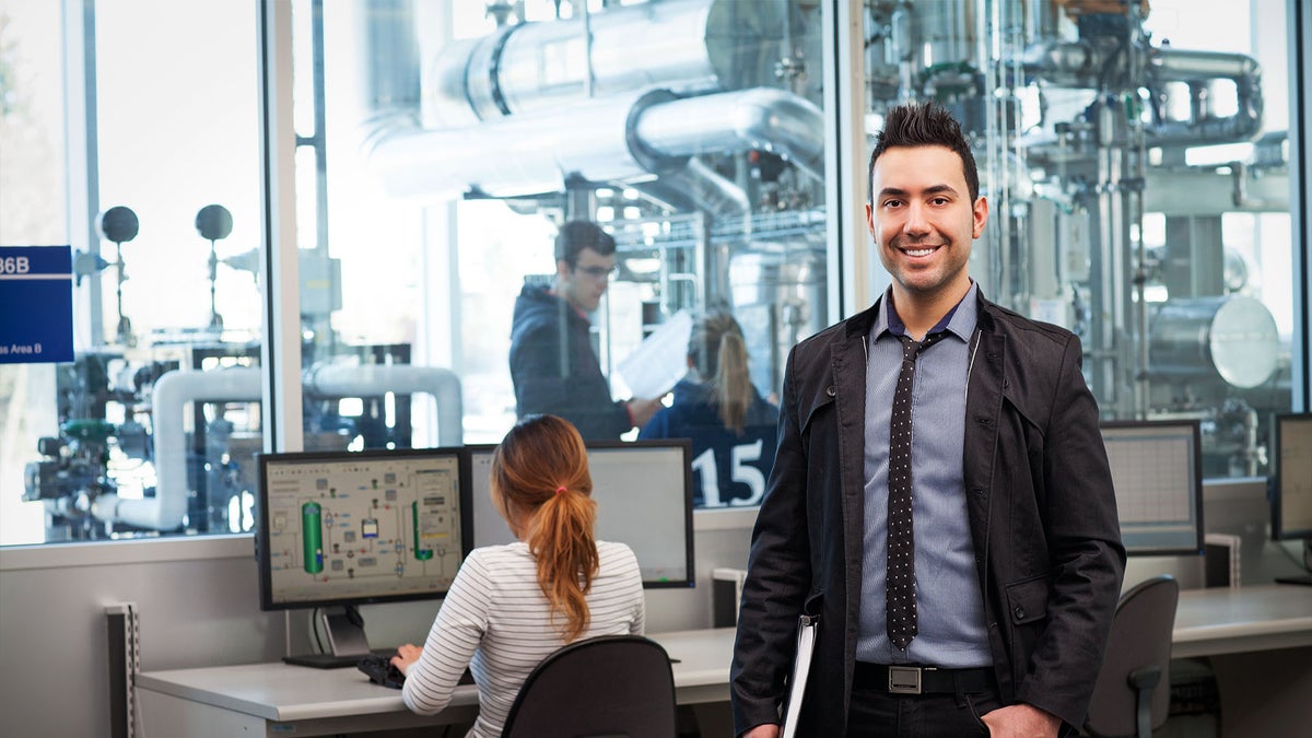 Man in gray suit in office smiling while holding a book, with woman at a desk on a computer in background.